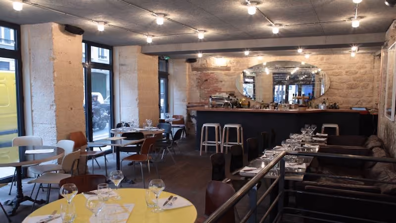 Interior of a restaurant with set tables, various chairs, stone wall, and a large oval mirror behind the bar.