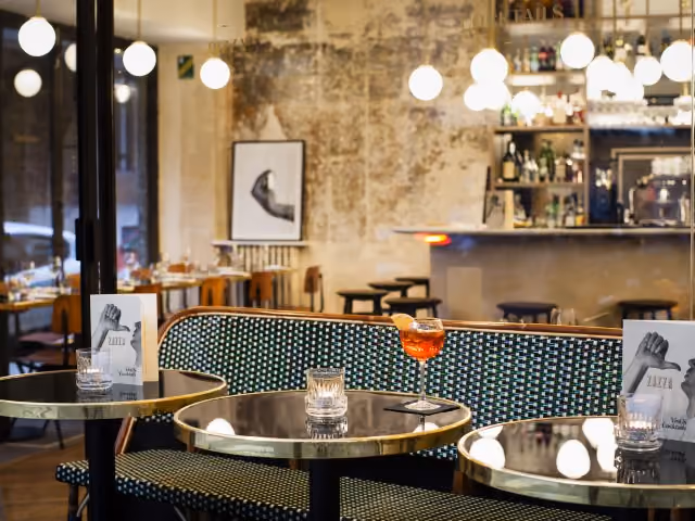 Interior of an elegant café with round brass tables, a green patterned sofa, an orange drink on a table, and a bar in the background.