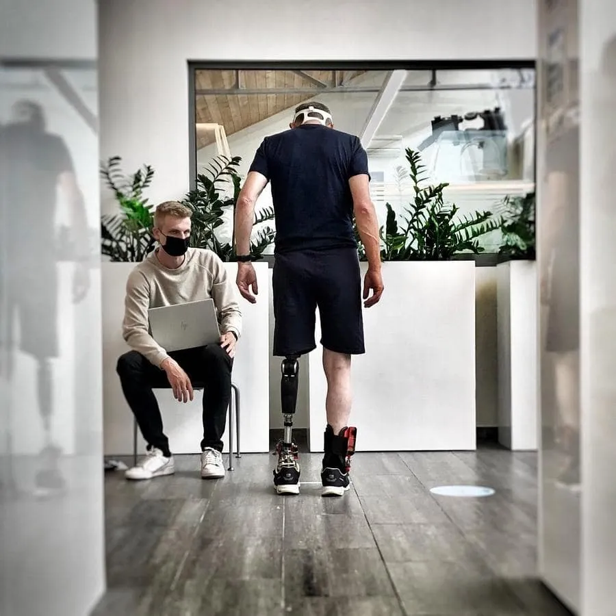 Man with a prosthetic leg walking indoors while another man wearing a mask sits nearby holding a laptop.