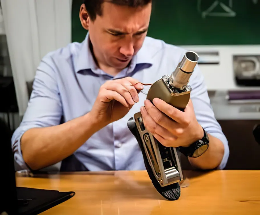 Man in light blue shirt examining and working on a prosthetic leg at a wooden table.
