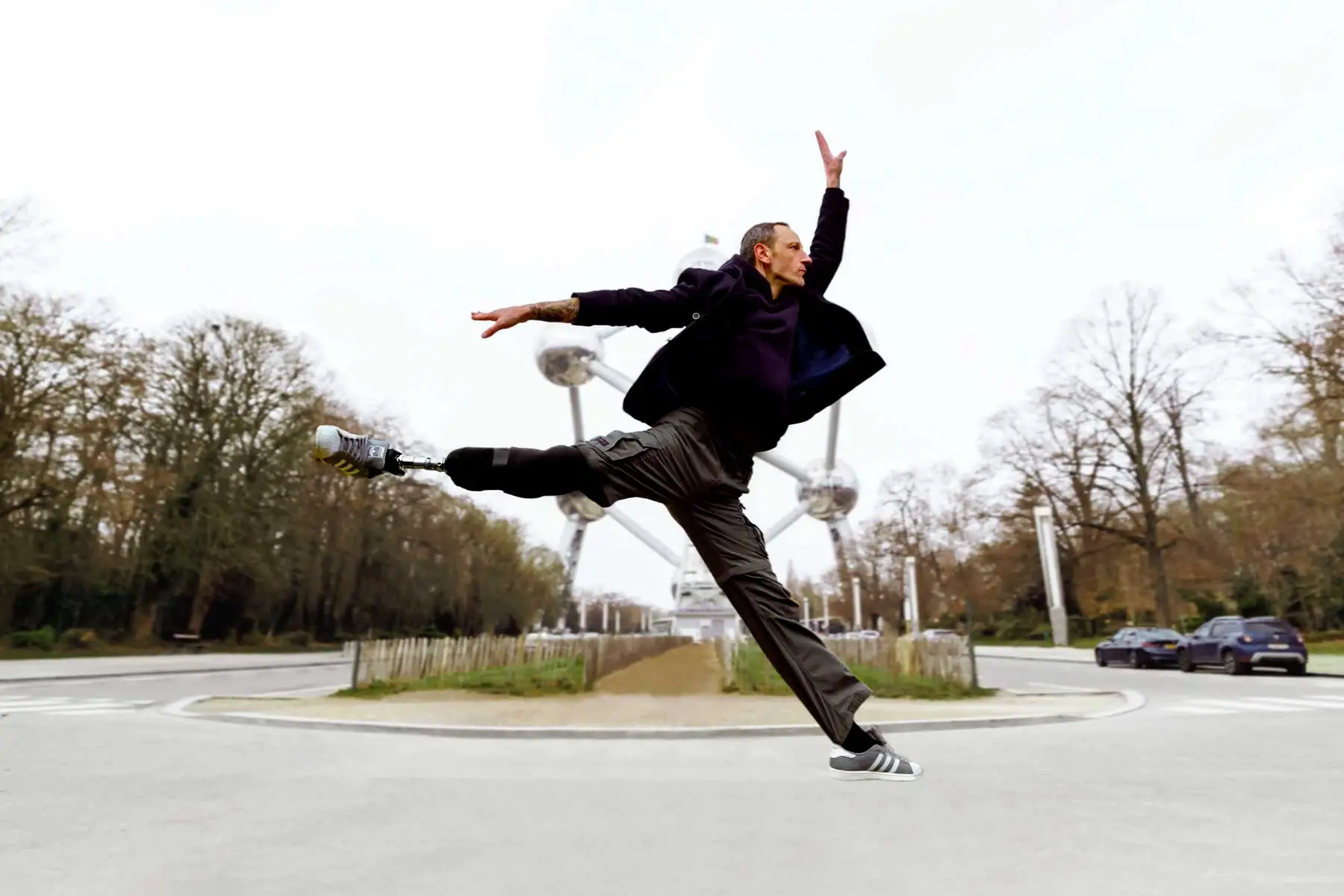 Man with a prosthetic leg jumping in an expressive dance pose outdoors with Atomium structure in the background.