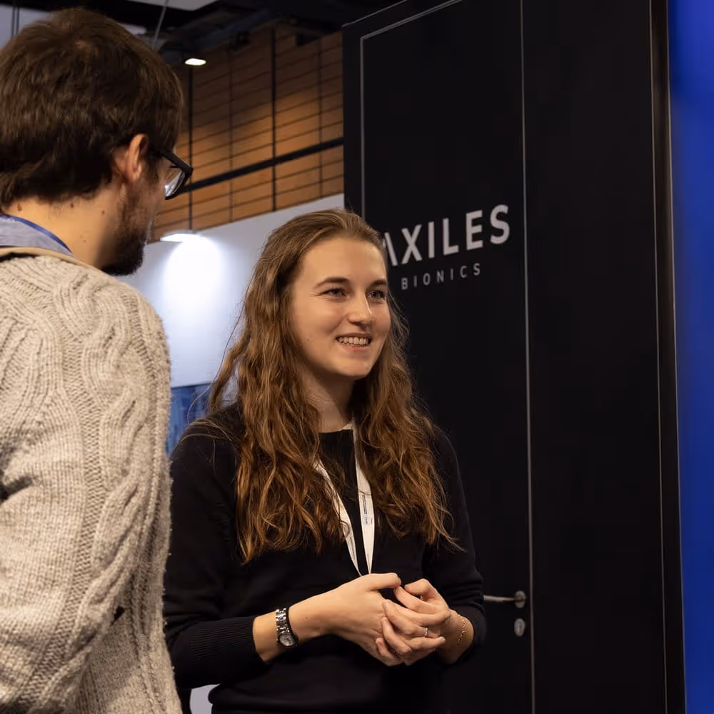 A woman with long hair smiling and talking to a man in a gray sweater at an Axiles Bionics booth in OT World.