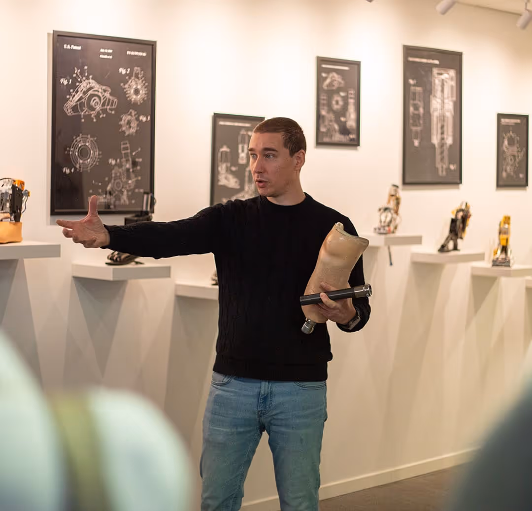 Man wearing black sweater and jeans explaining prosthetic legs in a museum with technical drawings and patents on the wall.