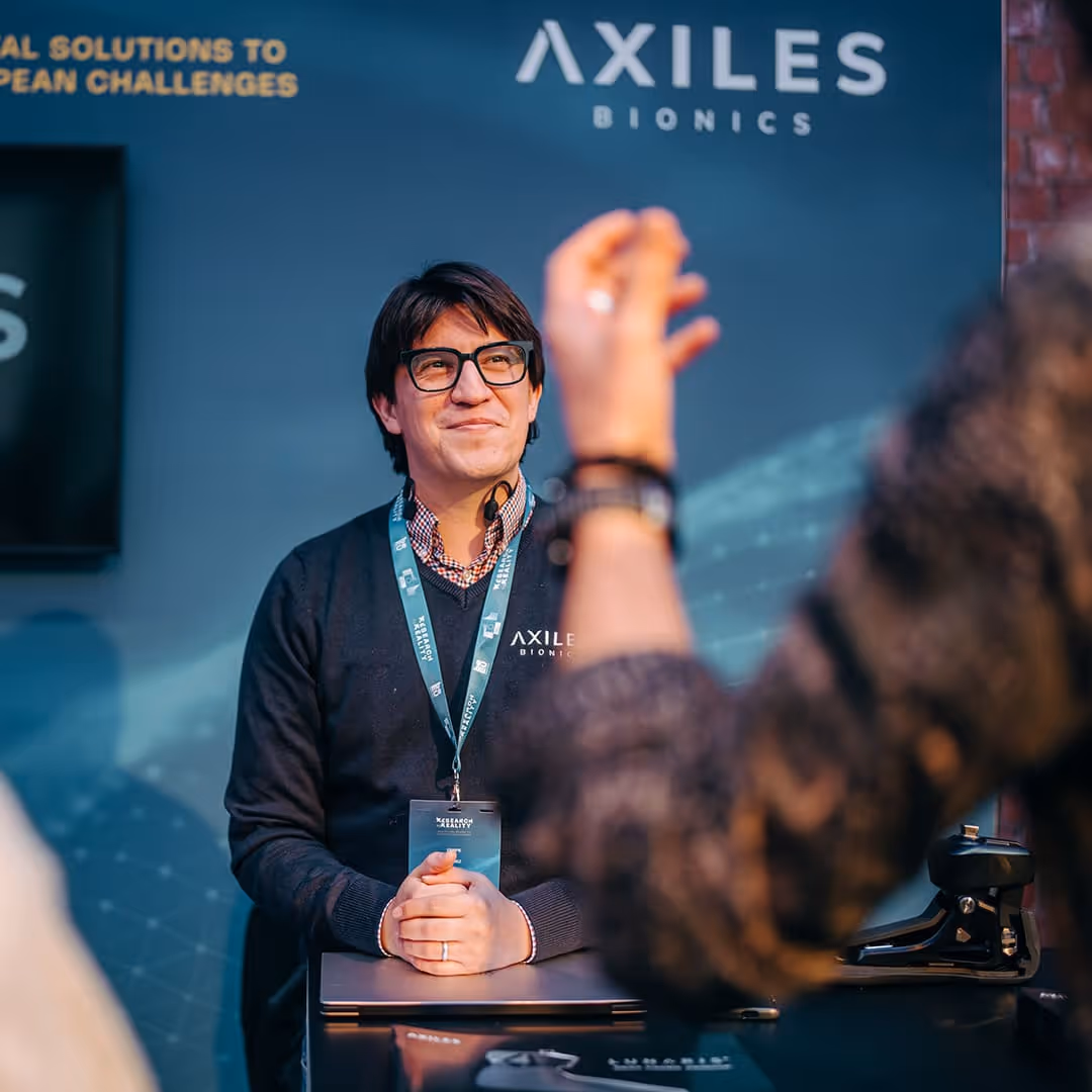 Felipe Gomez, CTO, with glasses and a lanyard sitting behind a desk at an Axiles Bionics booth, smiling and listening to someone gesturing in front of him.