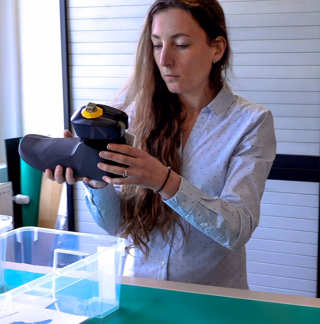 Claire Cherelle, CRO, with long hair holding and examining a black and yellow robotic or prosthetic foot indoors.