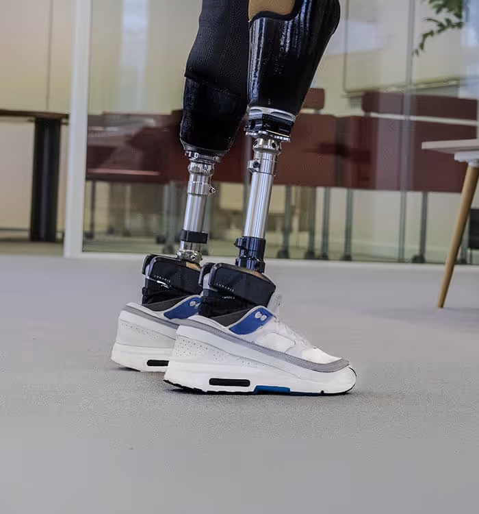 Close-up of double amputation bilateral Lunaris prosthetic legs with white and blue sneakers indoors on a gray floor.