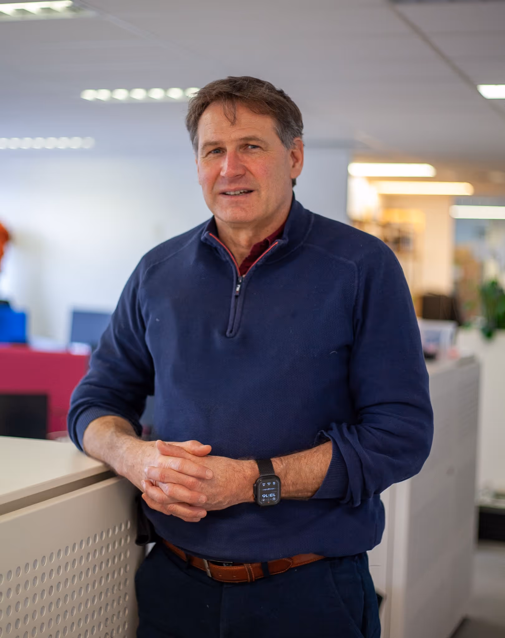 Jacques-Langhendries, CFO, in navy blue quarter-zip sweater leaning on a white office partition with hands clasped, wearing a smartwatch.