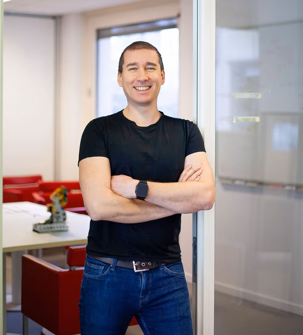 Smiling Pierre Cherlle, CEO of Axiles Bionics, in black t-shirt and blue jeans standing with arms crossed in an office near a glass door.