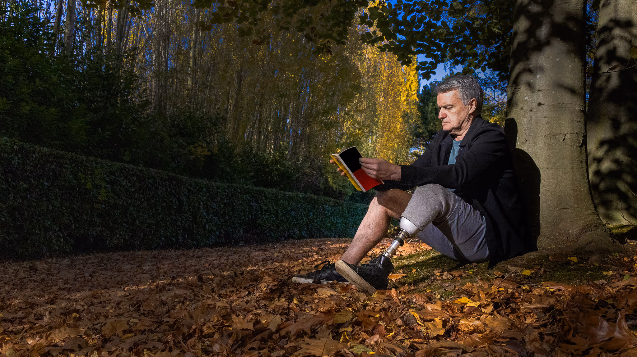Man with a prosthetic leg sitting under a tree reading a book on a ground covered with autumn leaves.