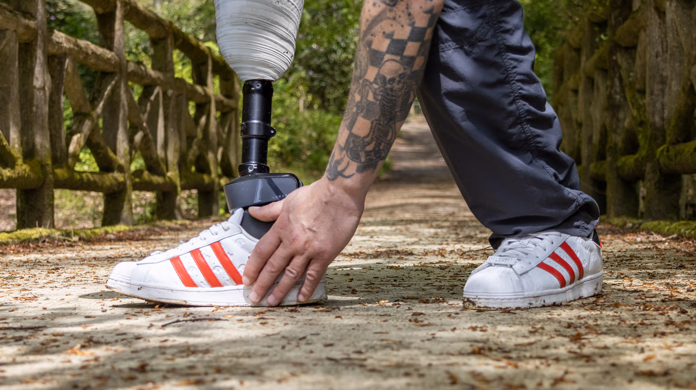 Person with a leg prosthesis wearing white shoes with red stripes, touching the shoe outdoors on a path with wooden railings.