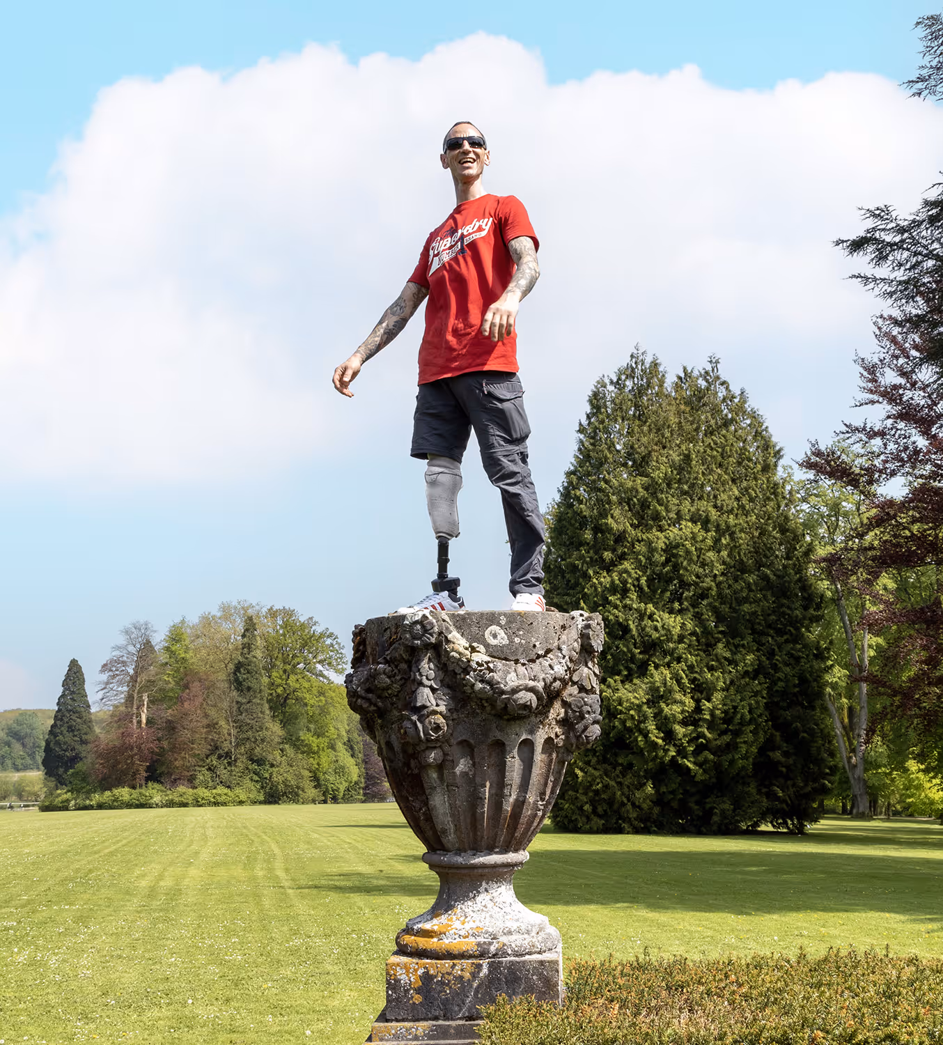 Smiling man with a prosthetic leg standing on a large ornate stone pedestal in a green park.