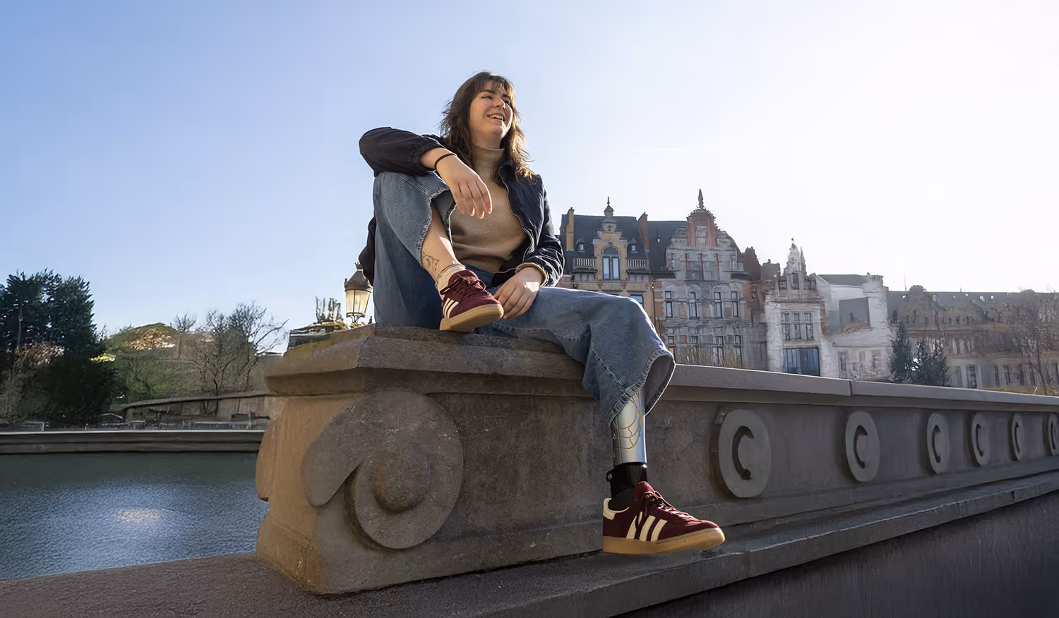 Smiling young woman with a prosthetic leg sitting on a stone railing by the water, in front of old European buildings under a clear sky.