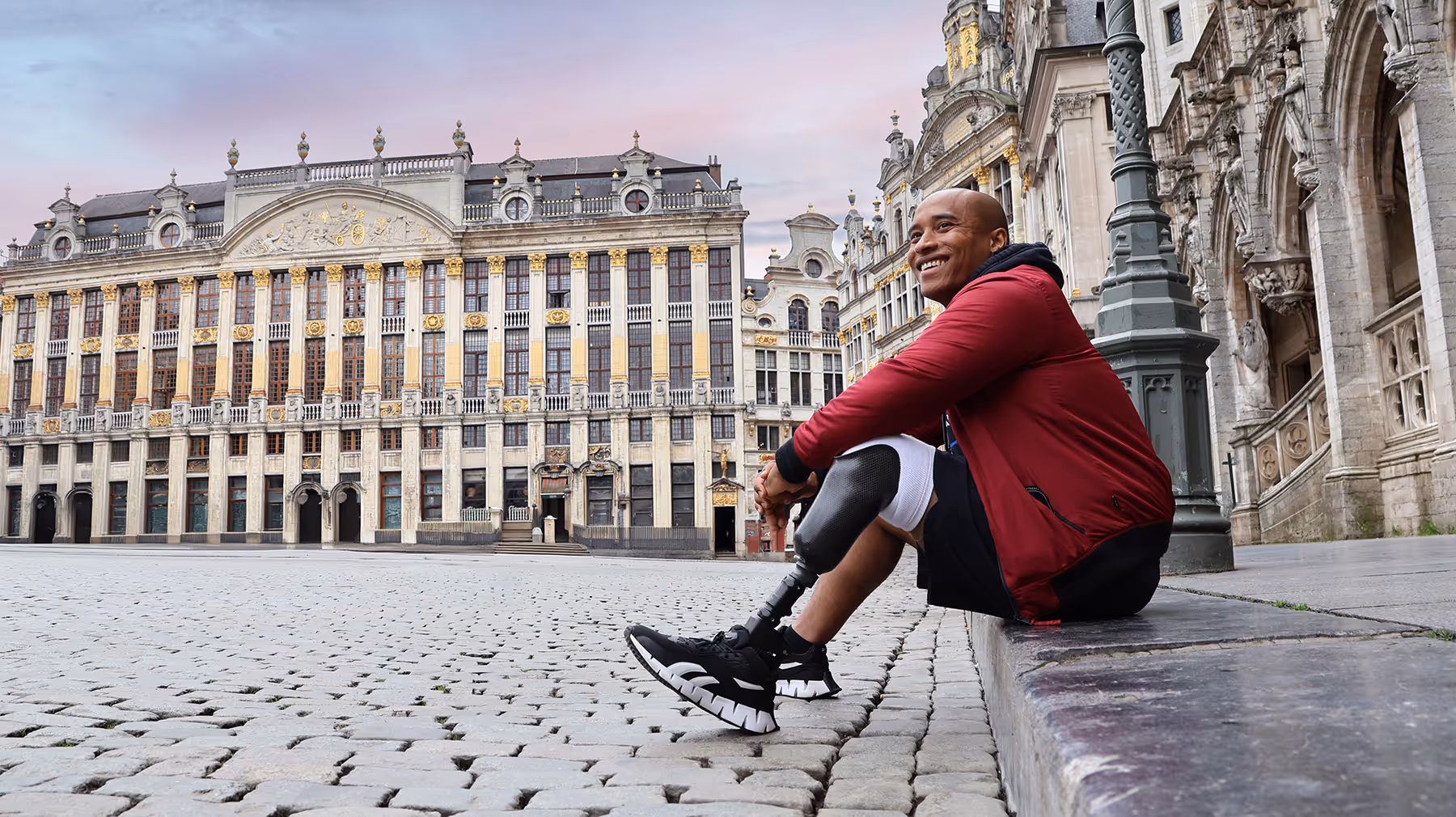 Smiling man with a prosthetic leg sitting on a stone ledge in a historic European square with ornate buildings.