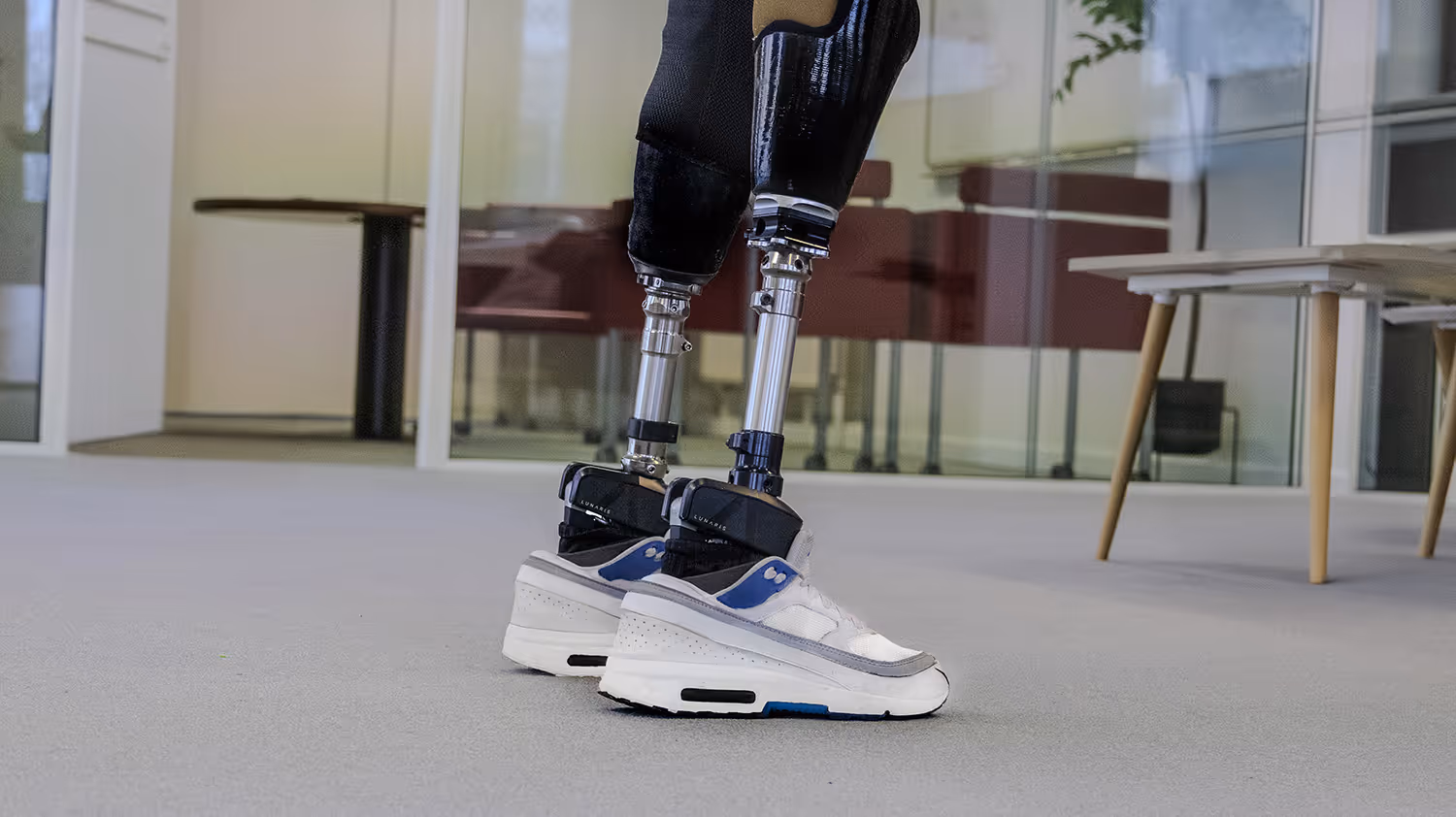 Close-up of a person wearing modern prosthetic legs with white and blue athletic shoes indoors.