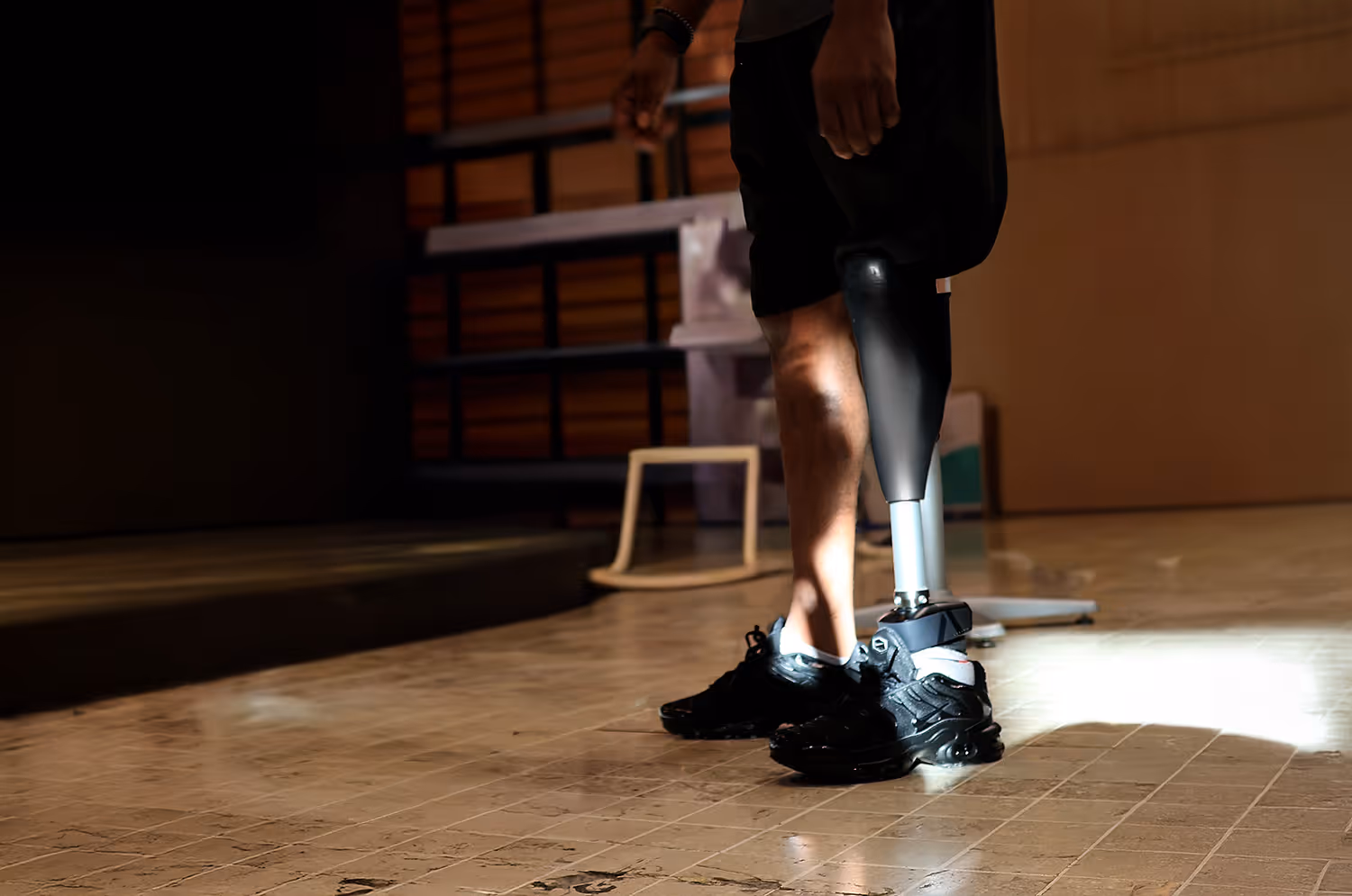 Person standing indoors on tiled floor wearing a black prosthetic leg and black athletic shoes.