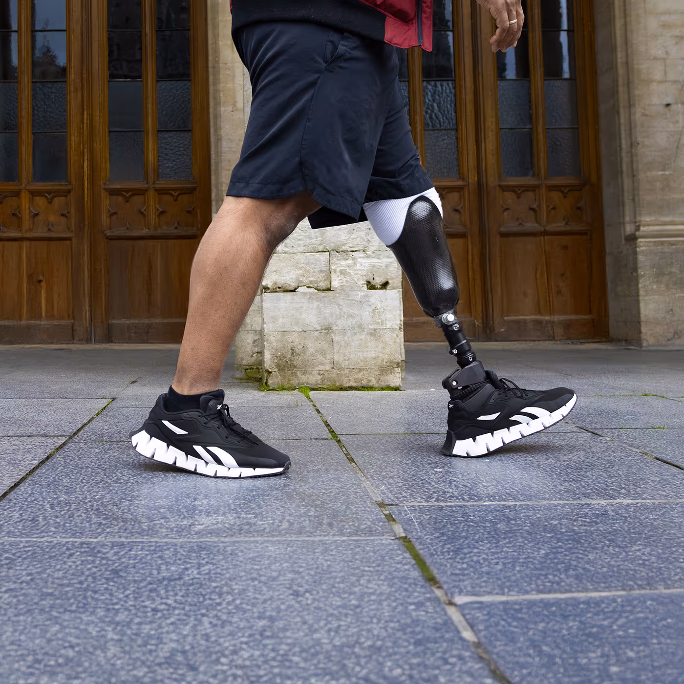 Person walking with a black prosthetic leg and wearing black and white athletic shoes on a tiled sidewalk.