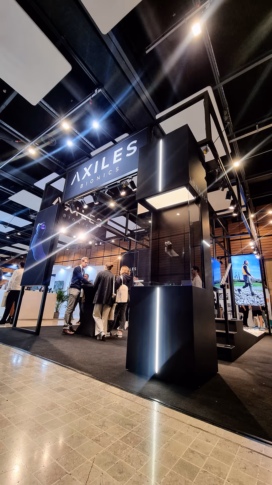 Trade show booth for Axiles Bionics with people interacting under illuminated signage and display cases labeled Lunaris.