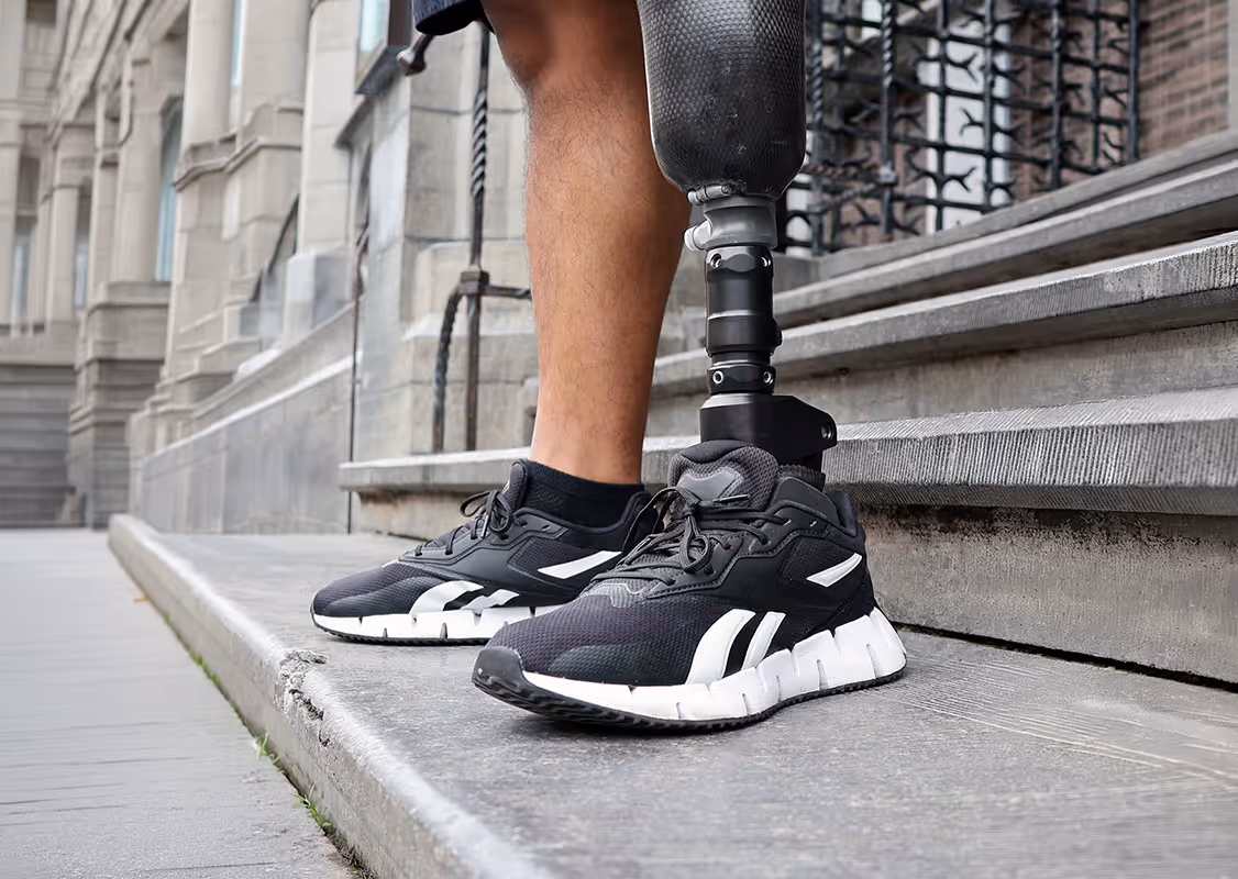 Close-up of a person wearing black sports shoes and a prosthetic leg standing on a stone step outside a building.