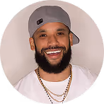 Smiling man with a beard wearing a gray cap, white shirt, and gold chains.