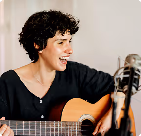 Young woman with curly hair smiling and playing an acoustic guitar near a microphone.
