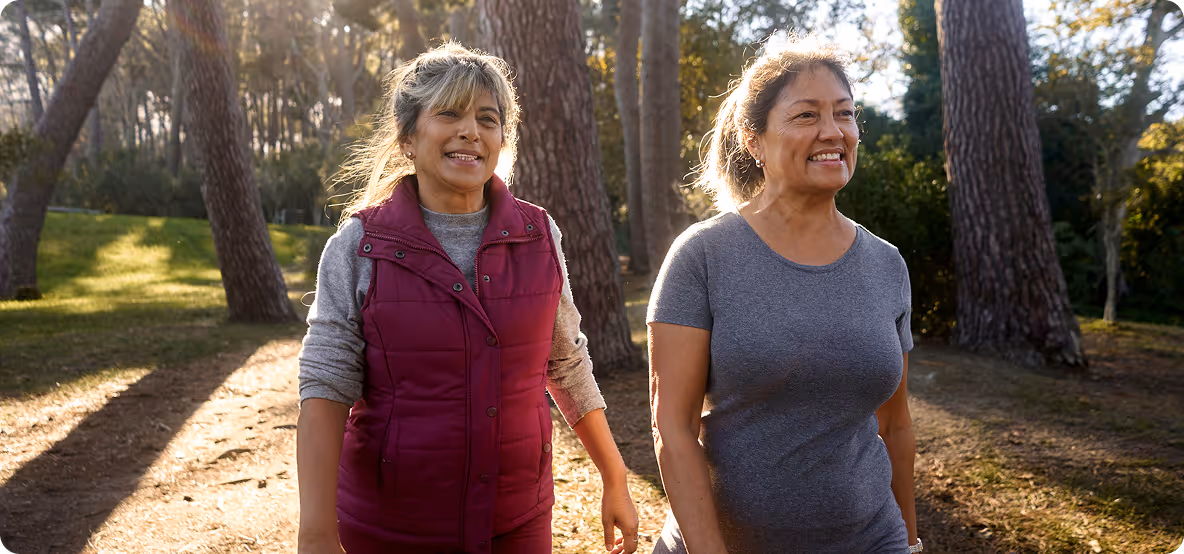 Two middle-aged women smiling and walking outdoors in a sunlit wooded park.