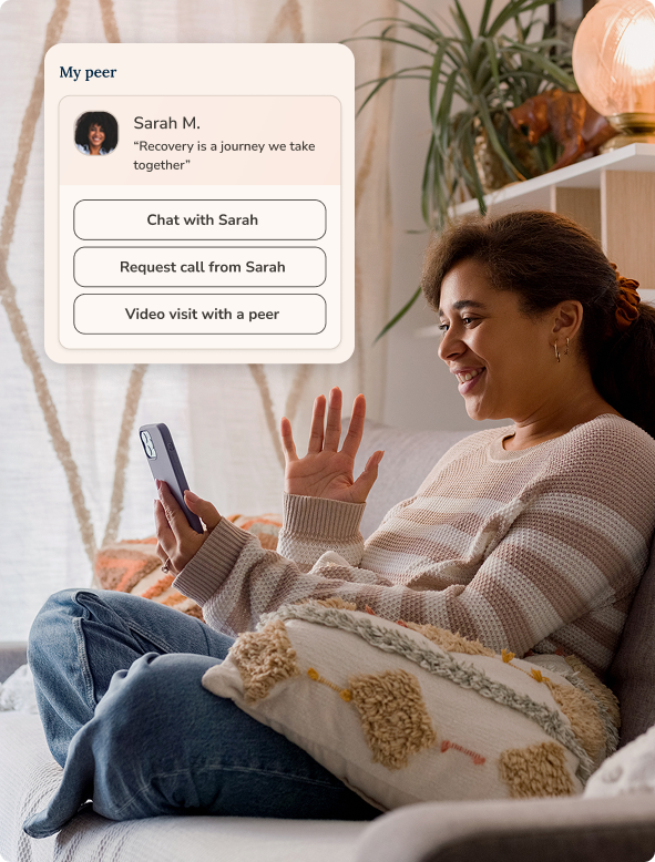 A woman sitting on a couch smiling and waving while holding a phone, with an overlay showing a chat app interface for connecting with a peer named Sarah M., including options to chat, request a call, or video visit.
