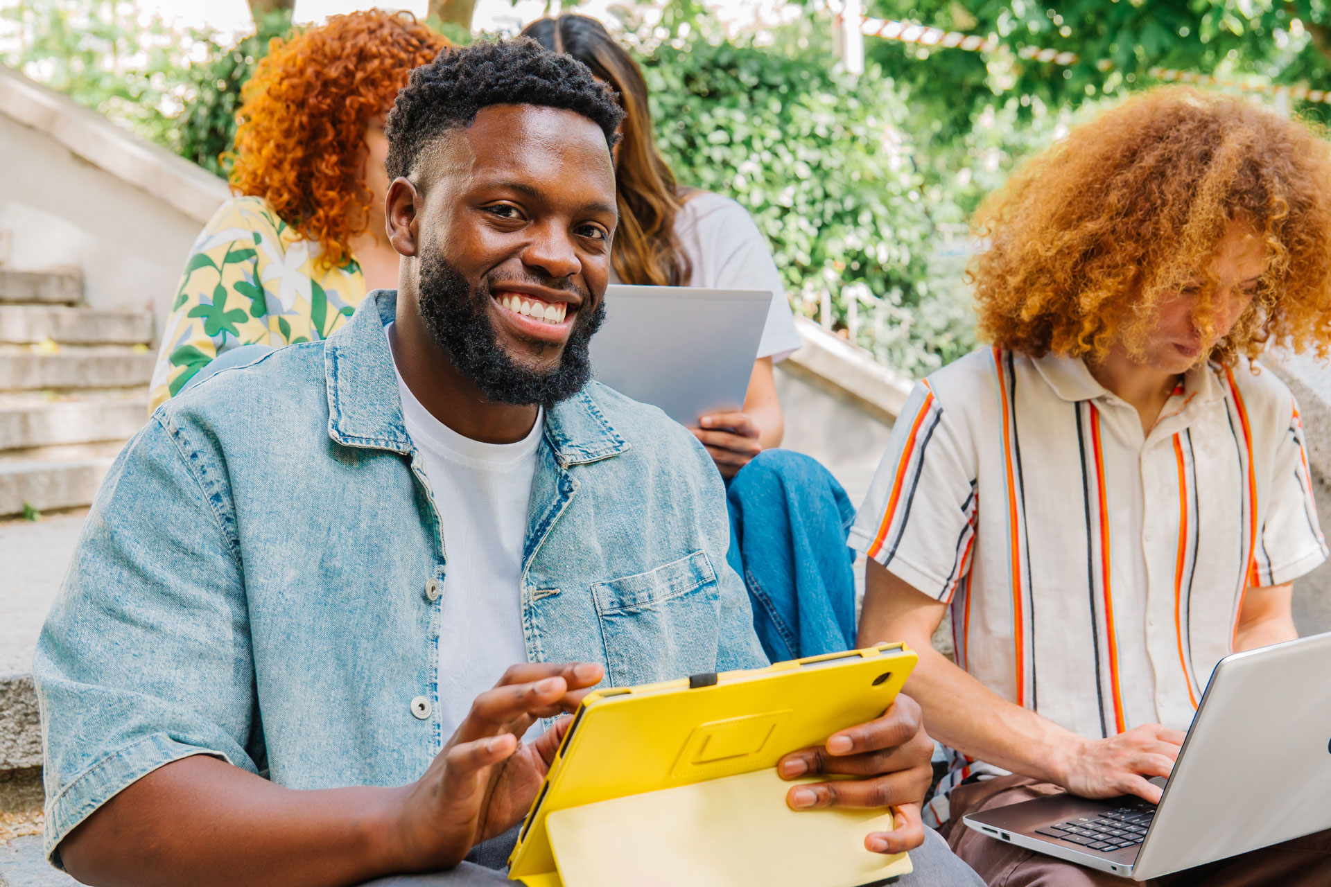 University students are using tablets and laptops while sitting on the stairs of their campus, enjoying the connectivity and convenience of modern technology for their studies.