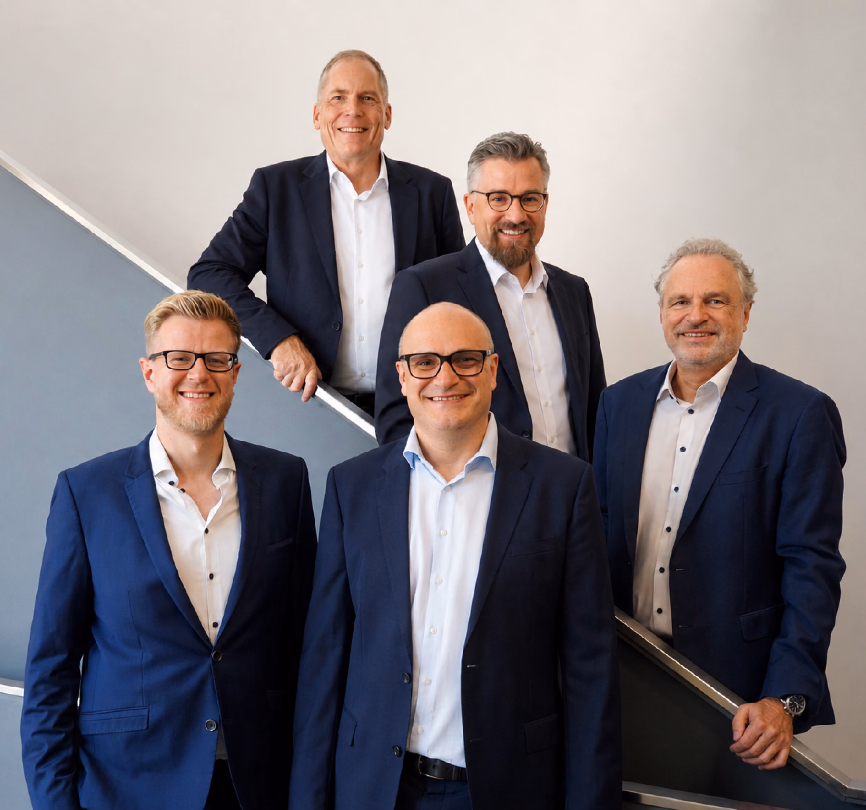 Five men in business suits standing on stairs smiling against a plain background.