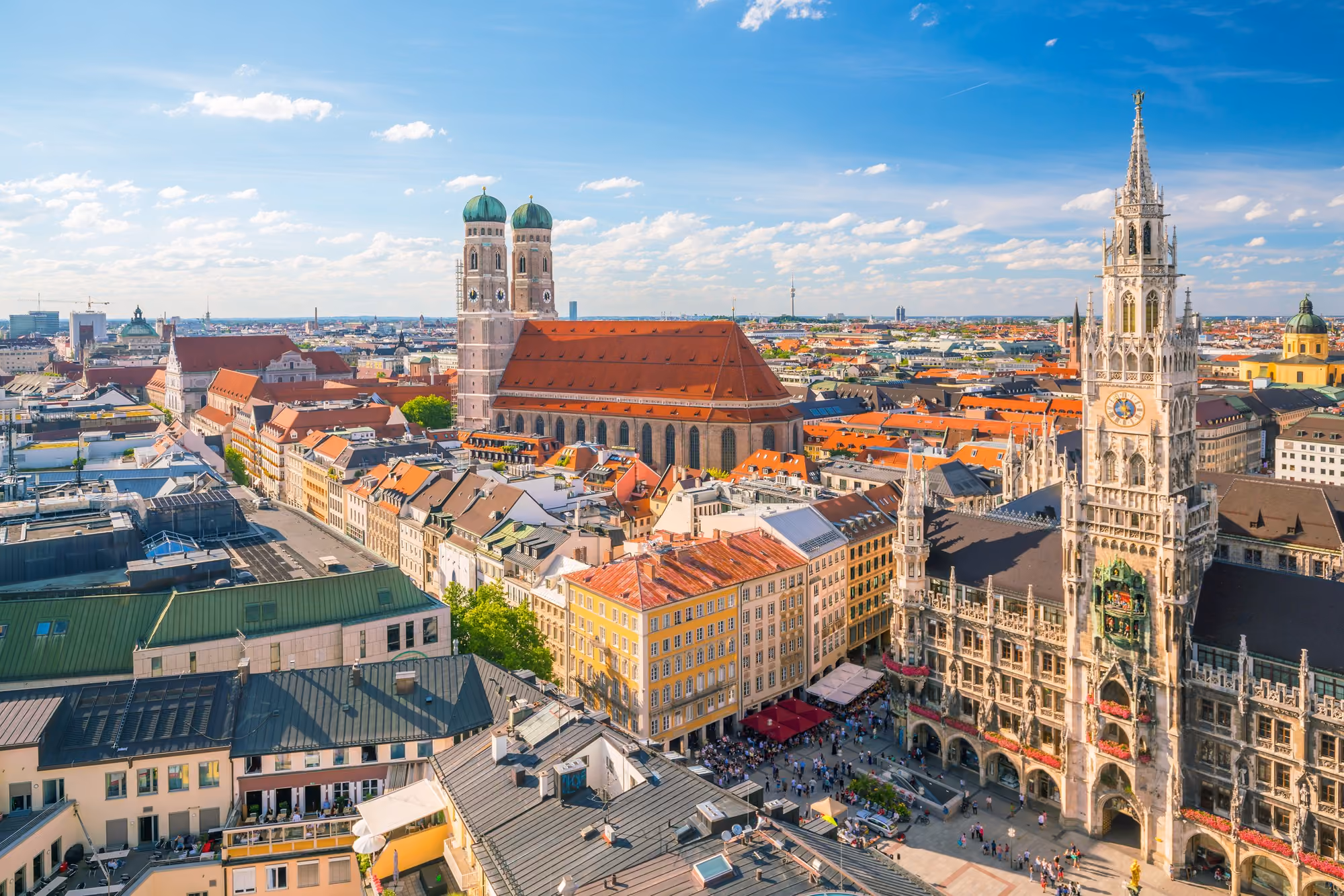 Aerial view of Munich, Germany, featuring the Frauenkirche cathedral with two green domed towers and the Neo-Gothic New Town Hall with its tall clock tower, surrounded by colorful rooftops and a busy town square under a partly cloudy blue sky.