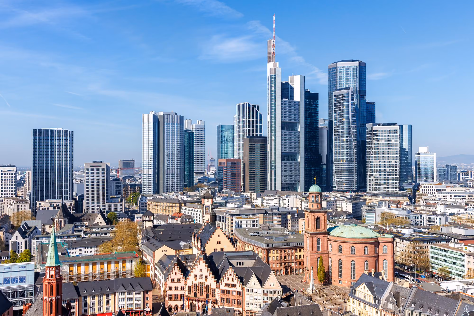 Cityscape of Frankfurt, Germany, featuring a mix of historic buildings with pointed roofs and a large red church with a green dome in the foreground, contrasted against a backdrop of modern glass skyscrapers under a clear blue sky.