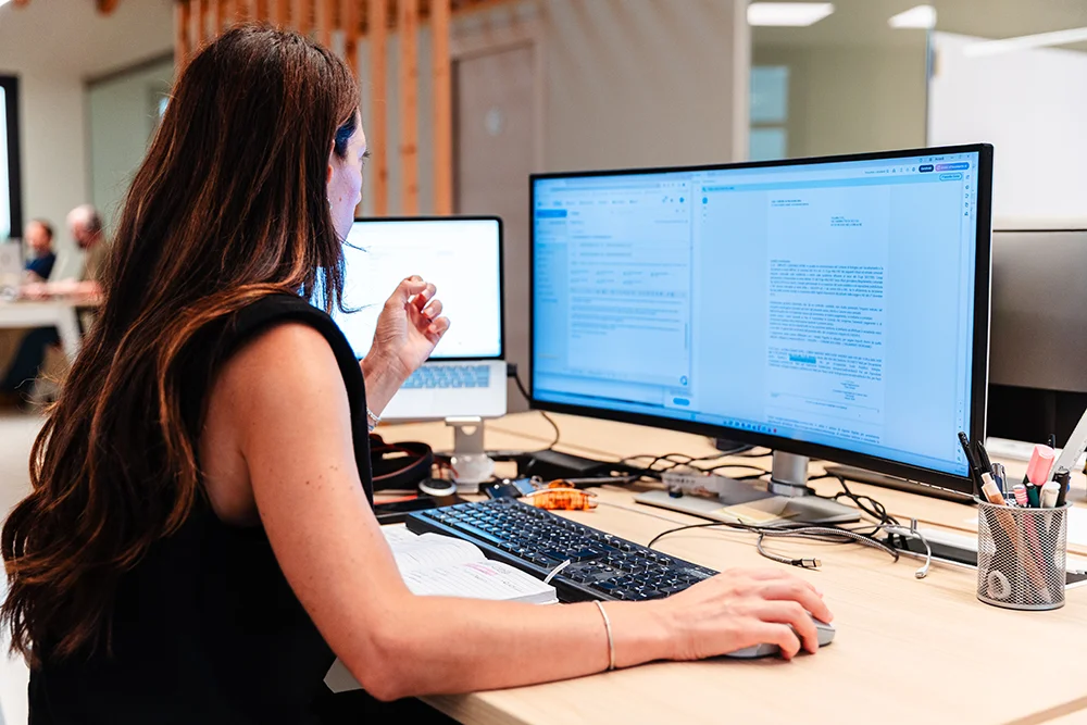 Donna al lavoro in un ufficio moderno, seduta a una scrivania con doppio schermo, che utilizza un mouse mentre guarda i documenti sul monitor principale.