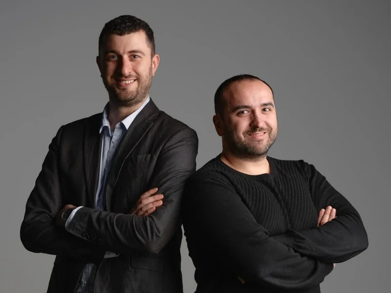 Due uomini sorridenti, uno con giacca nera e camicia azzurra chiara e l'altro con maglione nero, entrambi con le braccia incrociate davanti a uno sfondo grigio neutro.
