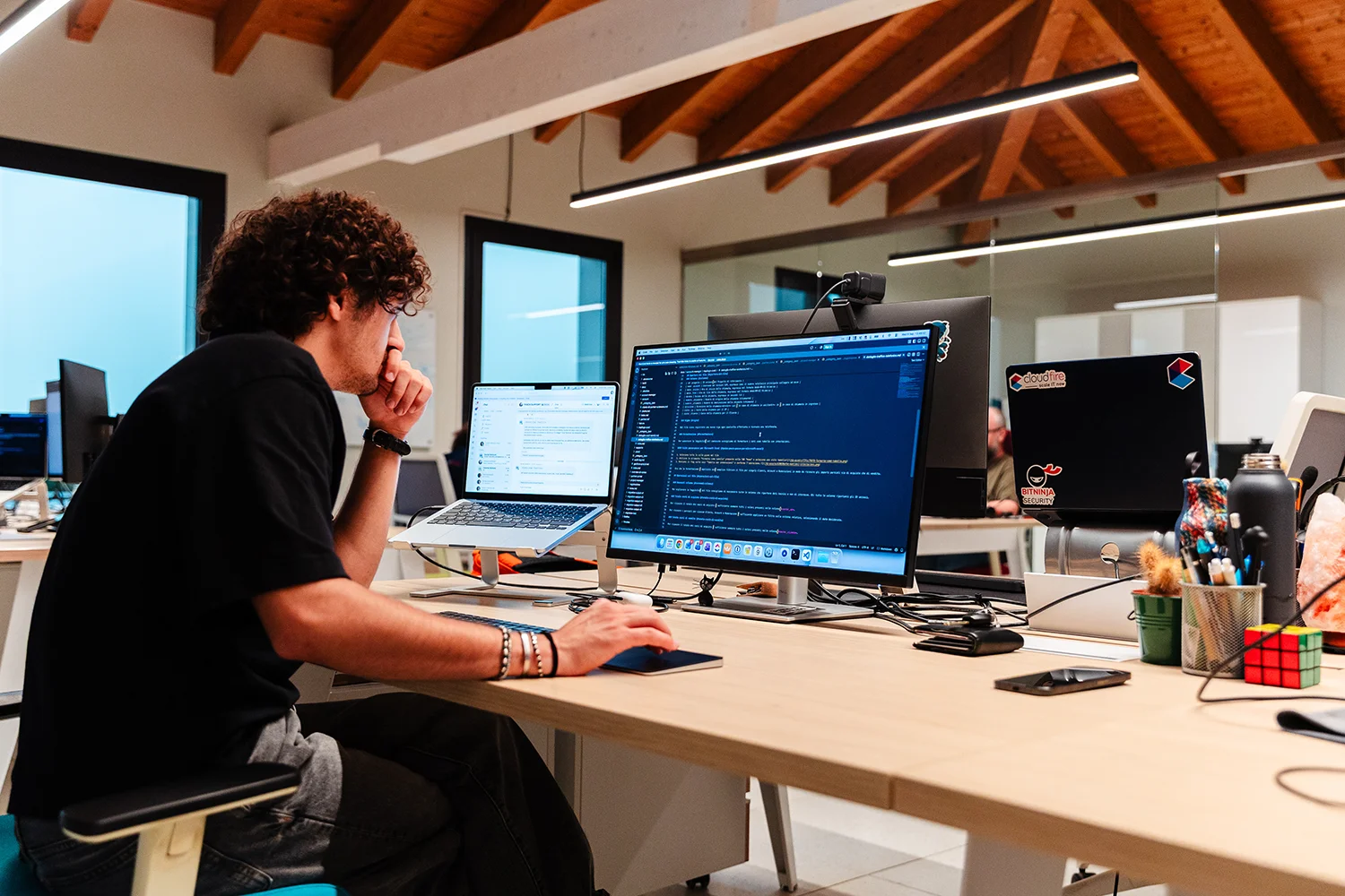 Uomo seduto a una scrivania in un ufficio moderno con soffitto in legno a vista, che lavora al computer con doppio monitor, uno mostra codice sorgente e l'altro messaggi di chat, con vari oggetti tra cui una tastiera, un mouse, una bottiglia termica, una pianta e un cubo di Rubik.