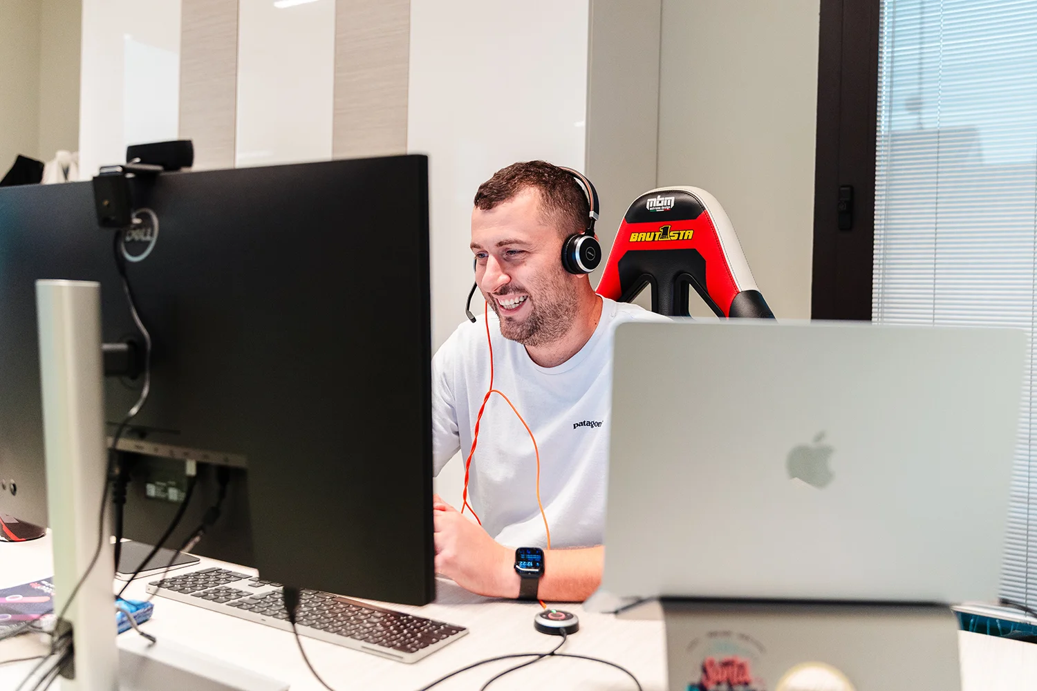 Roberto Bondavalli con cuffie e microfono che lavora al computer in un ufficio moderno, seduto su una sedia da gaming rossa e nera davanti a due monitor e un laptop Apple.