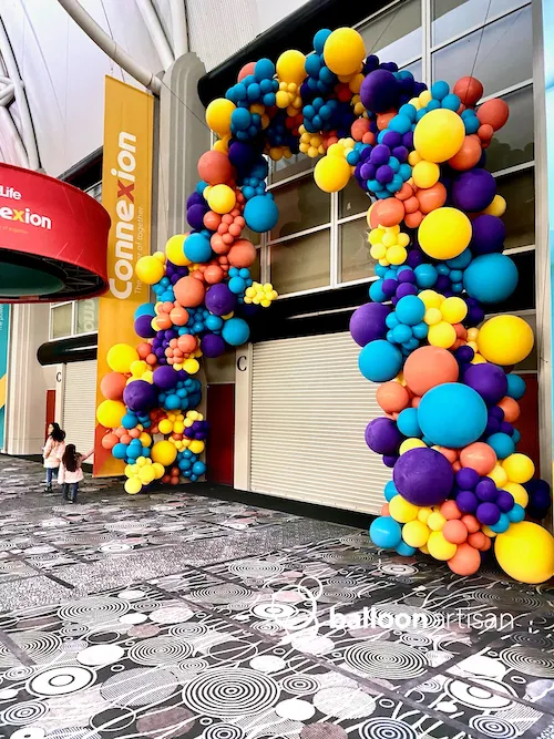 A large brightly colored balloon arch decoration for a balloon photo op at a convention hall spans over a large set of doors.
