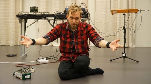 Tomas Hulenvik Klingberg, a composer, musician and workshop leader at ShareMusic, is in the picture. He sits on the floor with his hands up and conducts. He is dressed in a red- and black plaid shirt and black trousers. Behind him is a table with some musical instrument, next to the table is a theremin.