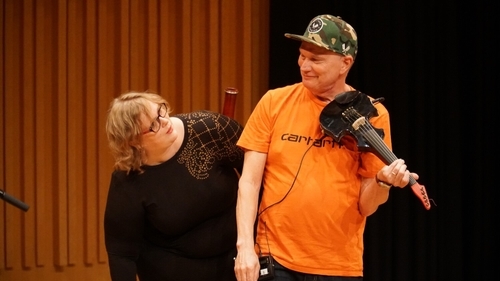 Two people are standing on stage close to each other. A woman with fair hair and glasses in a black shirt has stooped slightly and is looking curiously over her shoulder at a man. The man, wearing an orange shirt and a cap, is holding an electric black violin in his hands. He looks at the woman and smiles.