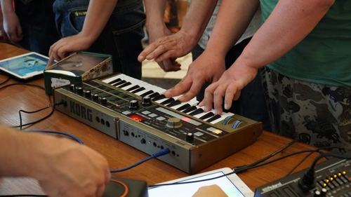 Some people play synthesizer together. The synth is on the table, centrally placed in the picture. There is a close-up of hands above and around the synth. Someone is pressing a key and someone is holding a cable.