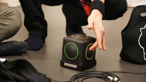 Some people are gathered around the musical instrument Skoog — a black cube with green, yellow and red circles on the sides. No faces are visible. A hand presses one of the circles.