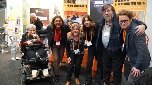 Gruppfoto på Folk och kultur 2020 vid ShareMusics monter. Sex personer står i led med händerna på varandras axlar och ler. Först till vänster syns en kvinna med grått hår i ShareMusics tröja och svarta byxor. Kvinnan sitter i en rullstol. Det är Lena Eldståhl, artist hos ShareMusic. Till höger om henne står Sophia Alexandersson, ShareMusics verksamhetschef och konstnärliga ledare. Sophia har svart lockigt hår och ett par glasögon och hon är kladd i en röd blus med rosett, svart kavaj och rutiga byxor. Till höger om Sophia står en kvinna med blont hår som är klädd  i en mörk blus och svarta byxor. Det är Linda Åkesson, bloggare och artist hos ShareMusic. Till höger om Linda står en tjej  med ljusbrunt hår som är klädd i svart. Det är Moa Johansson, teamledare på ShareMusic. Höger om Moa står en äldre man med skägg och glasögon. Han har en ljusblå skjorta, svarta byxor och en rutig halsduk. Mannen heter Nigel Osbourne, han är komprositör, professor och ordförande i ShareMusics akademiska råd. Till höger om honom står en yngre man i glasögon och med ljust lockigt hår. Han har på sig en grön kavaj och gråa byxor. Det är Richard Forsberg som var projektledare på ShareMusic.