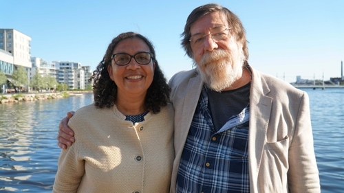 Sophia Alexandersson, ShareMusic's chief executive and Artistic Director, and composer Nigel Osbourne stand close together in an urban setting. Sophia is smiling, with curly hair and glasses, wearing a light-coloured shirt. Nigel has put his hand on Sophia's shoulder. He is wearing a grey shirt, a checkered shirt and a light jacket. Behind them is a lake and some buildings.
