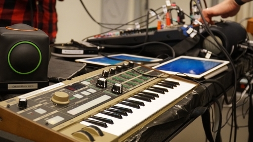 Some musical instruments can be seen in the picture. It's a synth, a mixer and two ipads. To the side is the musical instrument Skoog which looks like a black cube with one red and one green button on the sides.