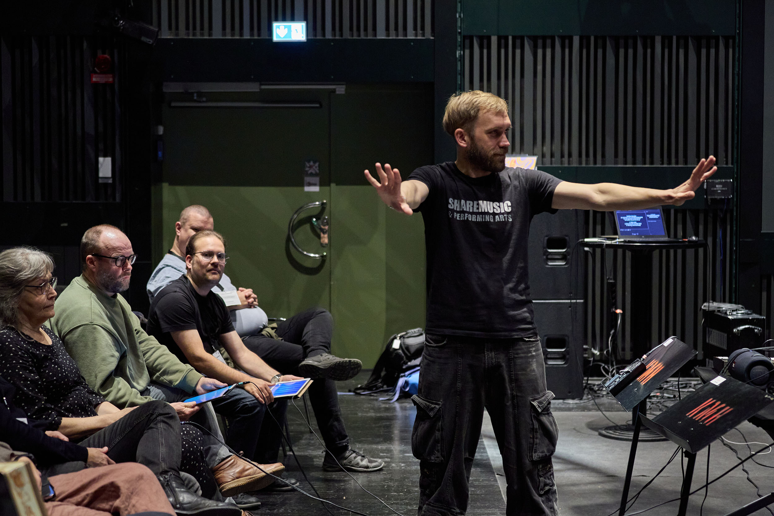 Bild från ett training lab i Skövde. Två personer, en man och en kvinna, står nära varandra. En kvinna med rött hår, glasögon och en brun tröja håller trumpinnar i händerna och ler. En man bakom henne är klädd i en blå tröja och har också trumpinnar. Han ser fokuserat in i kameran. Framför de står några mikrofoner. Bakom hänger en svart ridå.