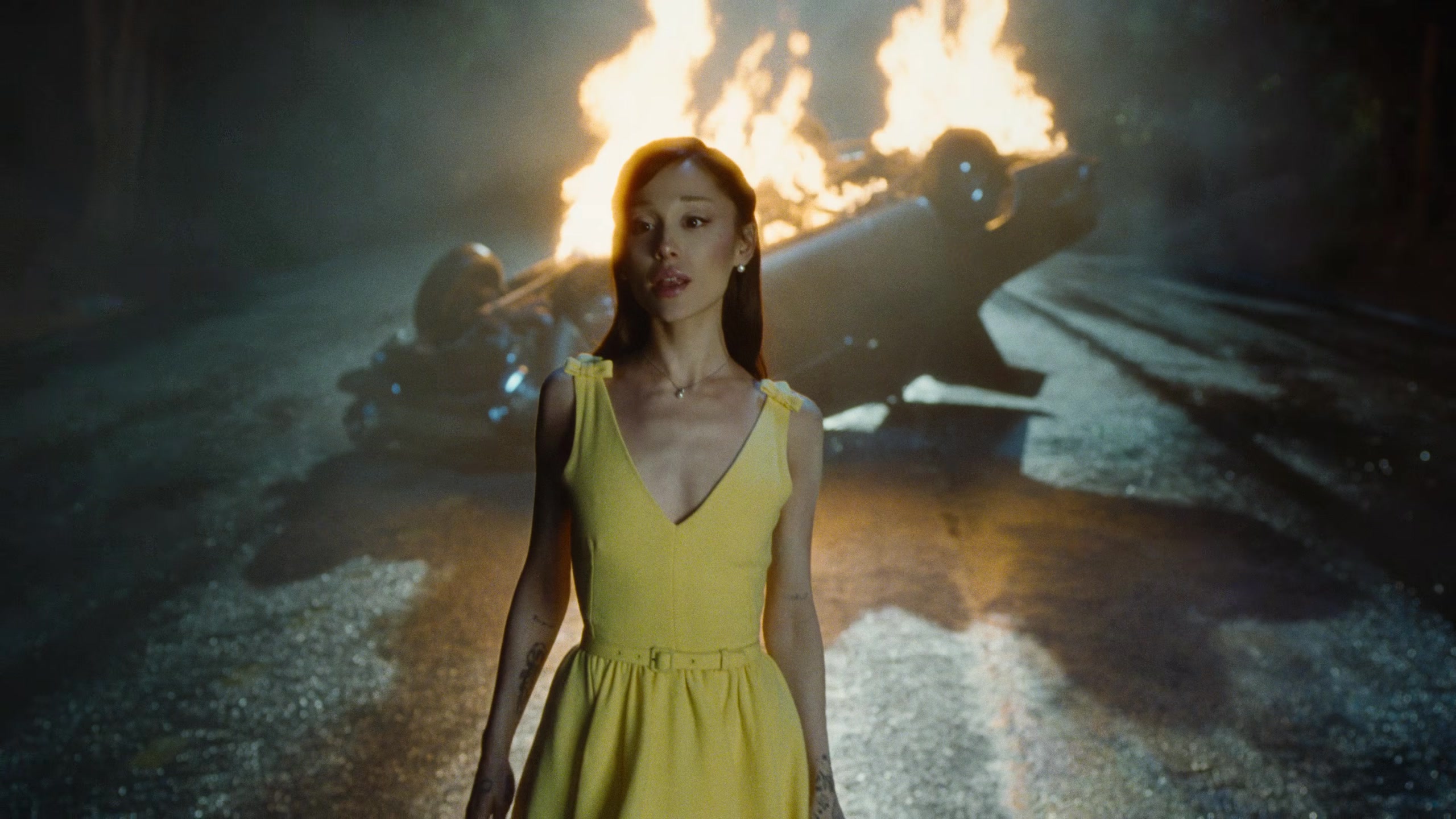 Ariana Grande in a yellow dress on a road at night, an overturned car burning behind her