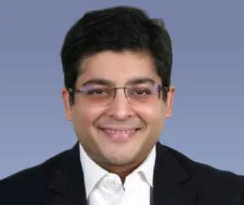 Smiling man with black hair and glasses wearing a white shirt and black blazer against a blue gradient background.
