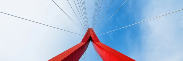 View looking up at a red cable-stayed bridge tower with cables fanning out against a blue sky with clouds.
