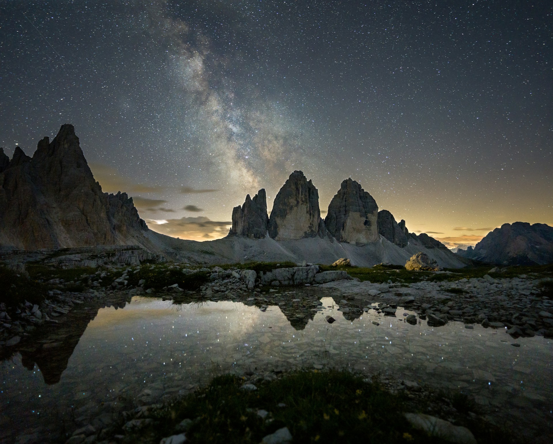 Night sky with stars and the Milky Way over jagged mountain peaks reflected in a still alpine lake.
