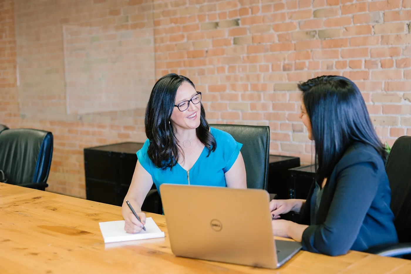 Two women smiling and talking at a table with a laptop and notebook in an office.