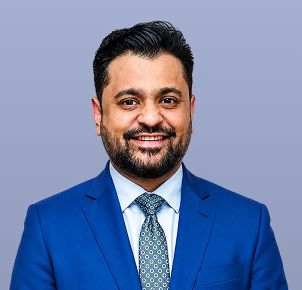 Smiling man with beard wearing a blue suit and patterned tie against a plain background.