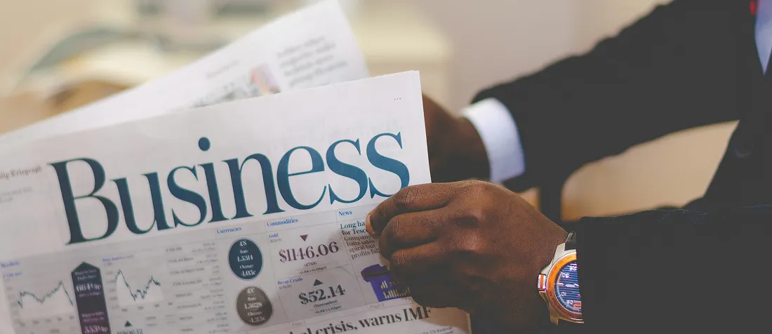 Person in suit reading a Business newspaper showing financial charts and market data.